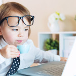 Smart toddler girl with glasses drinking coffee while using a laptop