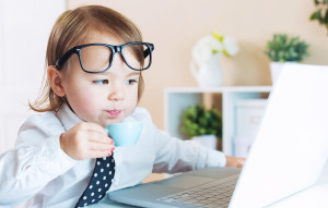 Smart toddler girl with glasses drinking coffee while using a laptop